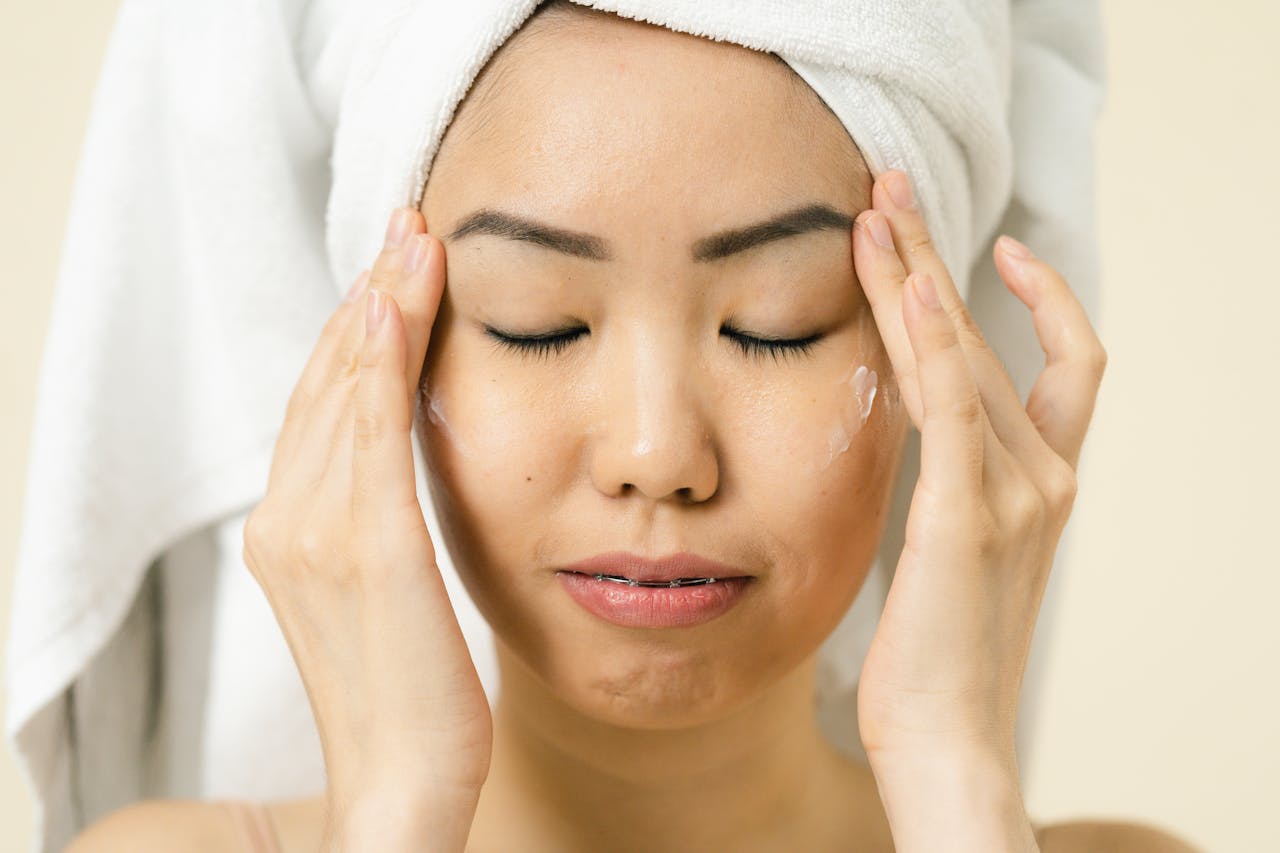 Asian woman with a towel on head applying moisturizer for skincare routine.
